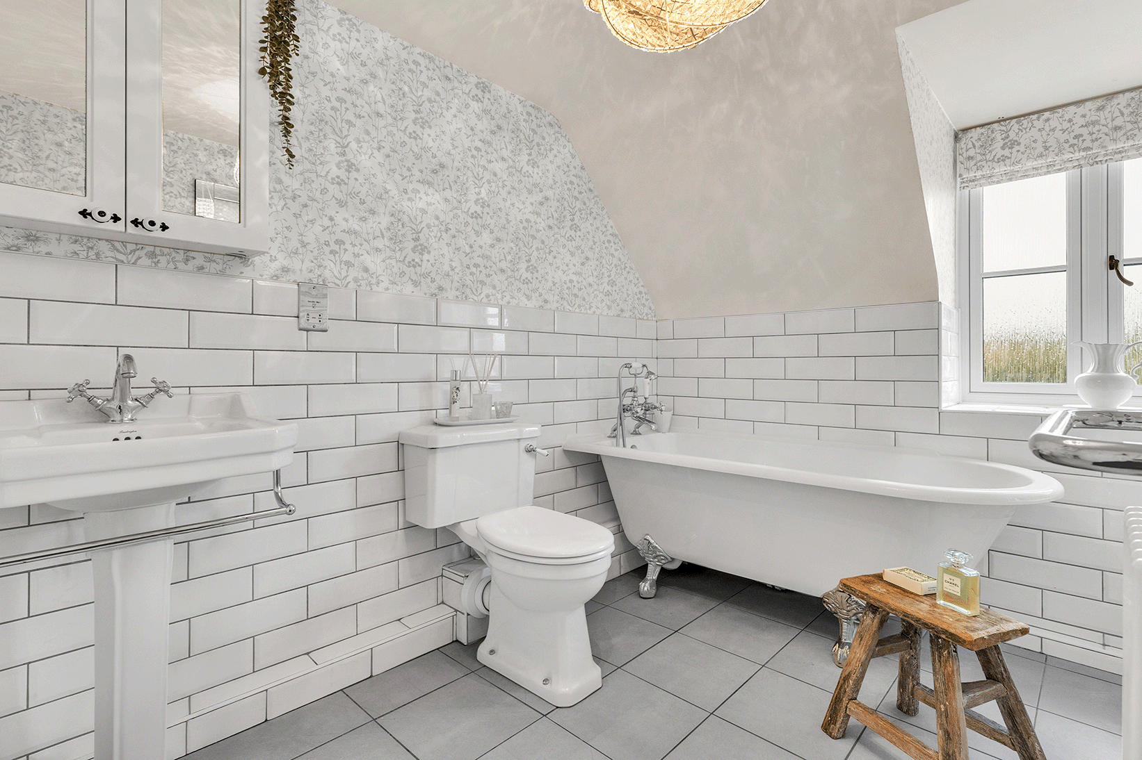 Elegant bathroom with freestanding clawfoot tub, pedestal sink, and white subway tile, featuring floral wallpaper and natural light, showcasing timeless design and buyer appeal in home upgrades.