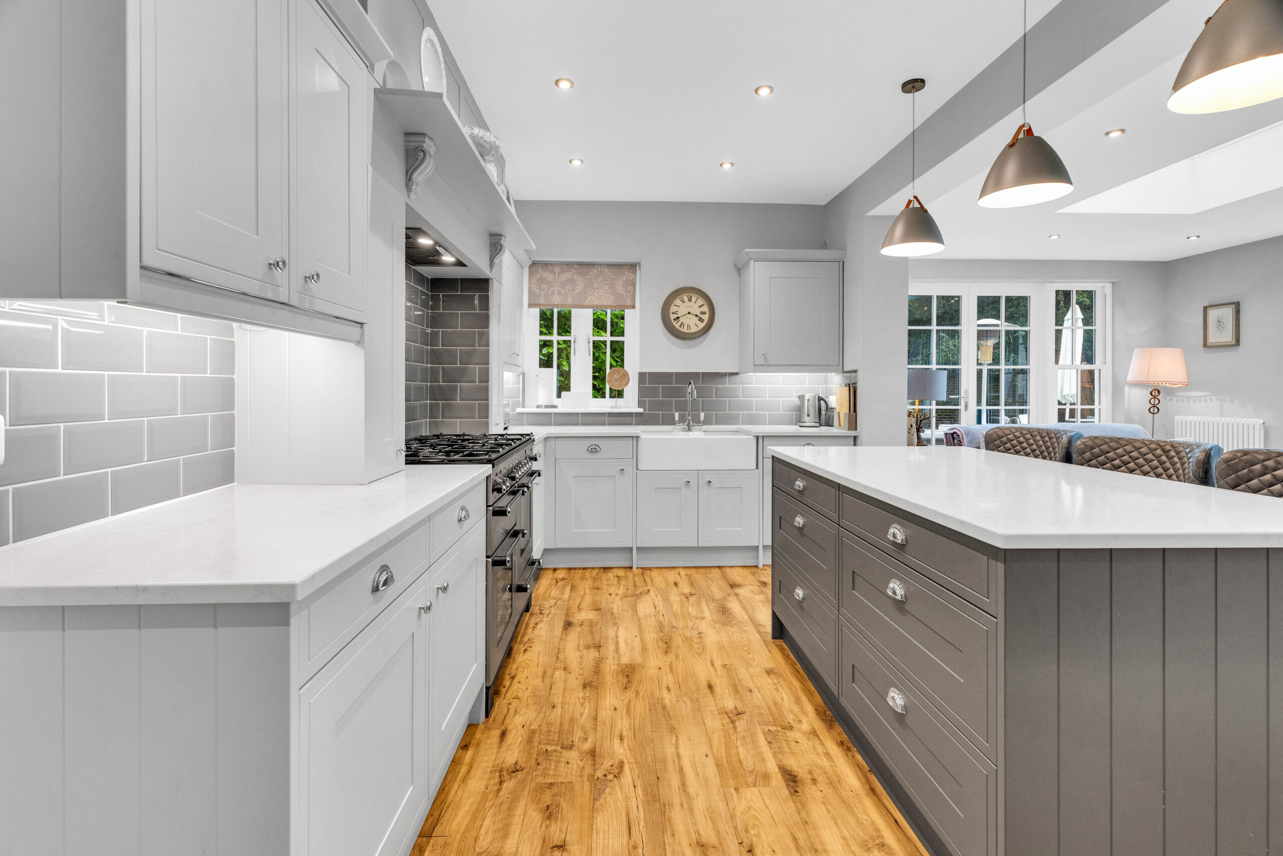 Modern kitchen featuring a large central island with shaker cabinets, white quartz countertops, grey subway tile backsplash, and natural wood flooring, with pendant lighting and a view into a cozy seating area.